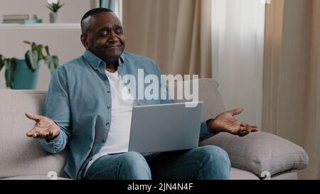 Reifer frustrierter afroamerikanischer Mann, der auf der Couch im Zimmer sitzt, schaut auf den Laptop-Bildschirm und liest schlechte Nachrichten in E-Mails, die frustriert sind Stockfoto