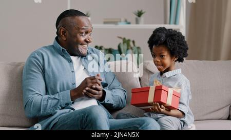 Glückliche kleine Tochter sitzt auf dem Sofa mit geschlossenen Augen Empfang Geschenkbox von liebevollen Vater fürsorglich Vater gratuliert Kind Mädchen Geburtstag Stockfoto