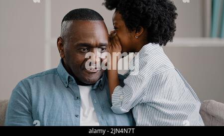 Kleine afroamerikanische Mädchen flüstert im Ohr enthüllt Geheimnis zu Papa Tochter teilt Geheimhaltung Klatsch vertrauliche Informationen reifer Vater sitzt auf Stockfoto