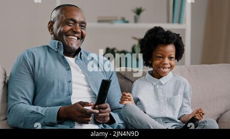 Nahaufnahme glücklicher afroamerikanischer Vater und Tochter, die lachen und sich einen lustigen fernsehfilm ansehen, der sich auf der Couch entspannt und die Fernbedienung mit dem Daumen hält Stockfoto