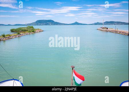 Ein Boot verlässt den Hafen von Fonyód am Plattensee in Ungarn. Winkende ungarische Flagge im Vordergrund. Stockfoto