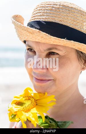 Frau in einem Strohhut mit Sonnenblume schaut zur Seite und wendet ihr Gesicht dem starken Wind, vor verschwommenem Hintergrund des Meeres. Stockfoto