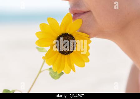 Nahaufnahme der unteren Hälfte des Gesichts einer Frau mit einer Sonnenblume vor einem verschwommenen Hintergrund des Meeres, Seitenansicht. Stockfoto