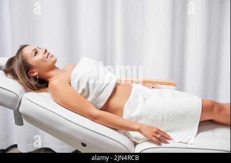 Fröhliche weibliche Klientin mit Handtüchern am Körper, die auf dem Tagesbett liegen und lächeln, während sie auf das Hautpflegeverfahren in der Kosmetologie-Klinik wartet. Fröhliche junge Frau mit perfekter Haut ruht im Schönheitssalon. Stockfoto