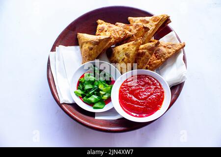 Samosa, samsa oder Somsa sind dreieckige frittierte Backwaren oder Pastri, die mit Tomatensauce und Porree-Scheibe auf schwarzem Hintergrund serviert werden Stockfoto