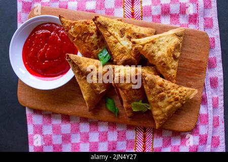 Samosa, samsa oder Somsa sind dreieckige frittierte Backwaren oder Pastri, die mit Tomatensauce und Porree-Scheibe auf schwarzem Hintergrund serviert werden Stockfoto