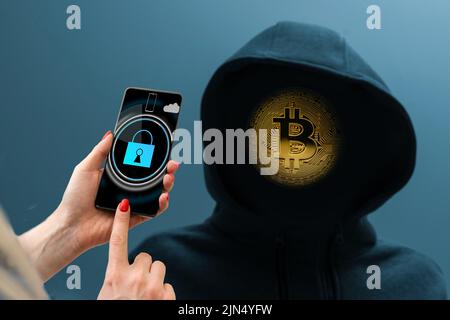 Ein Mann ohne Gesicht in einer Kapuze hält ein leuchtendes Bitcoin-Symbol auf einem dunklen Hintergrund. Stockfoto