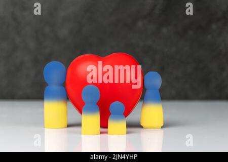 Vier kleine gelb-blaue Holzfiguren, die eine Familie symbolisieren - eine Mutter, ein Vater und zwei Kinder, die in der Nähe eines großen roten Herzens stehen. Das Konzept von f Stockfoto