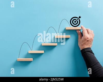Männliche Hand arrangiert eine hölzerne Blocktreppe mit Zielsymbol. Erreichen von Zielen oder Zielvorgaben. Stockfoto