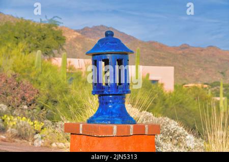 Blaue Säulenkopflampe gegen die Wüstenpflanzen und den Berg in Tucson, Arizona. Lampe auf einer braunroten Ziegelwand und einer Betonwand-Säule. Stockfoto