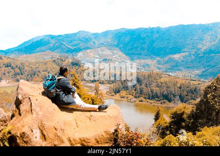 Mann, der auf einem Felsen einer Klippe steht und den Blick auf die Natur von See und Berg genießt. Symbol der Freiheit' Stockfoto