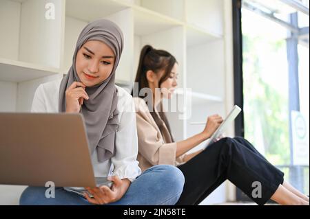 Die attraktive junge asiatische muslimische Frau nutzt einen Laptop, um ihre Hausaufgaben in der Bibliothek mit ihrer Freundin zu verwalten. Stockfoto