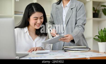 Glückliche und freundliche junge asiatische Geschäftsfrau oder Finanzarbeiterin, die mit ihrem Kollegen im Büro arbeitet. Stockfoto