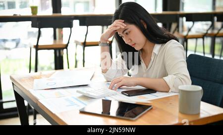 Junge asiatische Geschäftsfrau oder Buchhalterin, gestresst und nachdenklich, aus der Ferne von einem Café aus arbeitend und sich auf ihren Bericht konzentriert. Stockfoto