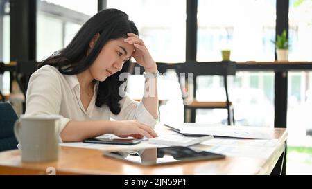 Gestresste und nachdenkliche junge asiatische Geschäftsfrau oder Buchhalterin, die im Café arbeitete und nachdenklich über ihren Verkaufsbericht nachdachte. Stockfoto