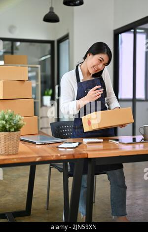 Beschäftigte und professionelle junge asiatische weibliche Online-Geschäft Inhaber bereitet ihre Versandkartons, während sie am Telefon im Büro sprechen. Stockfoto