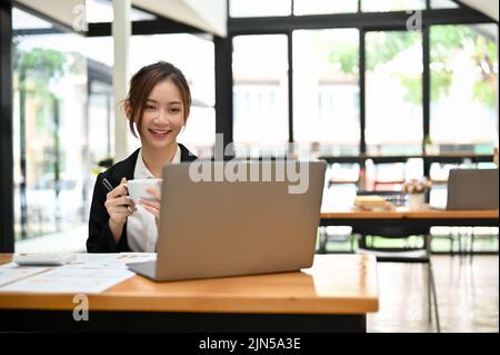 Attraktive junge asiatische Geschäftsfrau oder weibliche Büroangestellte, die im Coffee Shop arbeitet, Kaffee getrunken und ihre Aufgaben auf ihrem Portabl erledigt Stockfoto
