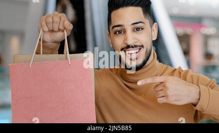 Glücklicher junger arabischer Käufer, der lächelnd im Einkaufszentrum steht und Einkaufstaschen in der Hand hält und mit dem Zeigefinger auf Geschenkpakete zeigt, die sich über Verkaufsrabatte freuen Stockfoto