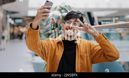 Junger glücklicher indischer Kerl blogger hält Smartphone-Aufzeichnung vlog Dreharbeiten Video für Blog in Mall fröhlichen Mann winken Gruß in Webcam auf Handy-Video Stockfoto