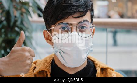 Nahaufnahme Porträt junger Mann, der drinnen steht und auf die Kamera posiert, trägt eine medizinische Einweg-Maske und zeigt den Daumen nach oben Stockfoto