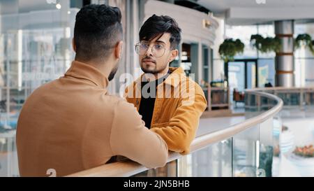 Zwei junge positive indische Jungs stehen in der Lobby und reden über Geschäftsproblem sorglose Freunde diskutieren über den Austausch von Nachrichten Freundschaft genießen Stockfoto