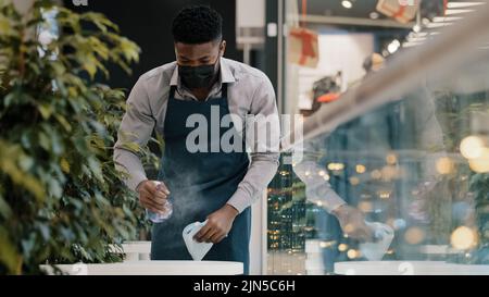 Junger männlicher Kellner in Schürze wischt Tisch Sprays Desinfektionsmittel Reiniger bereitet Café-Restaurant für die Eröffnung ernsthafte afroamerikanische Student in Medizin Stockfoto