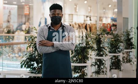 Kellner Barista Barkeeper Restaurantmitarbeiter Verkäufer trägt medizinische Maske posiert drinnen erfolgreiche selbstbewusste junge Café-Besitzer Geschäftsmann steht in Stockfoto