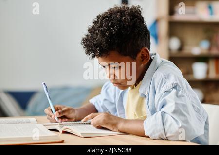 Afrikanischer Schuljunge, der im Zimmer an seinem Schreibtisch sitzt und Hausaufgaben macht, Notizen in Notizbüchern macht Stockfoto