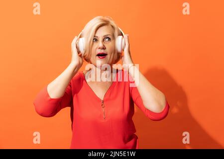 Schockierte Frau mittleren Alters mit blonden Haaren in Kopfhörern isoliert auf orangefarbenem Hintergrund. Konzept der Emotionen, Mimik Stockfoto