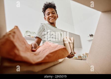 Blick aus der unteren Perspektive auf einen afrikanischen Jungen, der Papier in einer Pappschachtel zum Recycling wirft, während er in der Küche steht Stockfoto