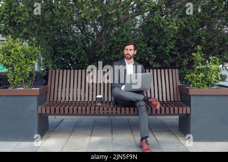 Erfolgreicher männlicher Geschäftsmann, der mittags auf einem Laptop auf einer Bank in der Nähe des Büros sitzt Stockfoto