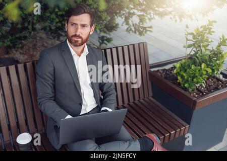 Erfolgreicher Geschäftsmann, der mittags auf einem Laptop arbeitet und auf einer Bank in der Nähe des Büros sitzt und wegschaut Stockfoto