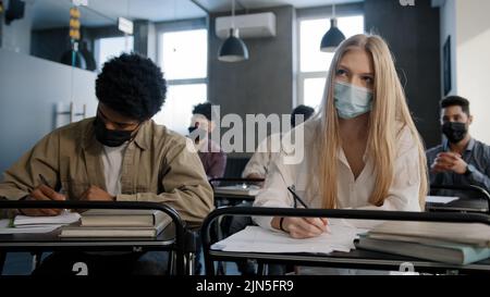 Verschiedene Studenten in medizinischen Maske sitzen im Klassenzimmer in der Lektion konzentriert hören Vortrag schriftlich Notizen in Notizbuch junge Menschen ernsthafte Schüler Stockfoto