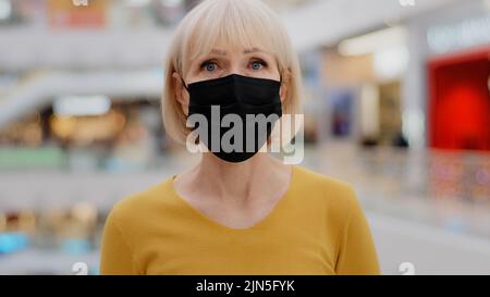 Nahaufnahme reife kaukasische Frau in Schutzmaske stehend drinnen Blick auf die Kamera besorgt ernst bewusst elegante Dame in medizinischen Atemschutzgerät Stockfoto