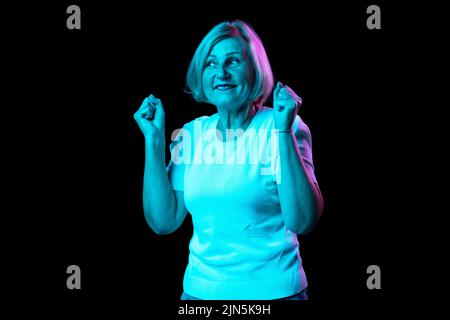 Halblanges Porträt einer kaukasischen älteren Frau mit blondem Haar, das isoliert auf dunklem Hintergrund in Neonlicht posiert. Konzept der Emotionen, Gesichtsbehandlung Stockfoto