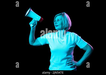 Halblanges Porträt einer kaukasischen älteren Frau mit blondem Haarstandinbg mit Megaphon auf dunklem Hintergrund in Neonlicht isoliert. Stockfoto