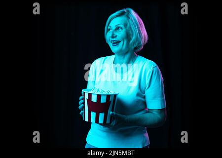Eine aufgeregte ältere Frau, moderne Großmutter, die einen Eimer Popcorn auf dunklem Hintergrund in Neon hält. Konzept der Emotionen, Gesichtsausdruck Stockfoto