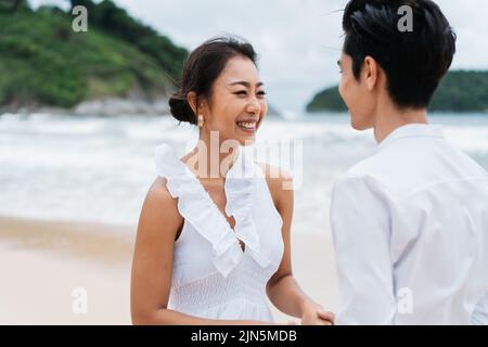 Glücklich toothy lächelnd Asian 30s Paar in Beziehung in offen am Strand im Sommer. Ein romantisches Paar in einem weißen Hemd und Kleid. Stockfoto