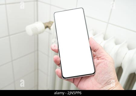 Modell, Hand hält ein Smartphone mit blanko weißem Display vor einem Heizkörper, Konzept für steigende Kosten aufgrund von Energiekrise und Inflation Stockfoto