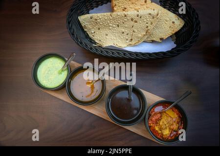 Indische Dip, Relish, Chutney und Sauce in kleinen schwarzen Schalen mit knusprigem Papadam-Brot auf einem dunklen Holztisch serviert, Kopierer Platz, Blick von einem Stockfoto