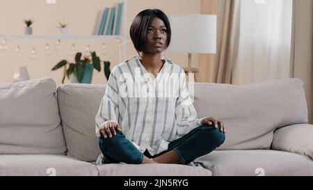 Junge fokussierte afroamerikanische Frau, die in Lotusposition mit geschlossenen Augen sitzt gesundes ruhiges Mädchen, das sich auf dem Sofa entspannt und tief durchatmen kann Stockfoto