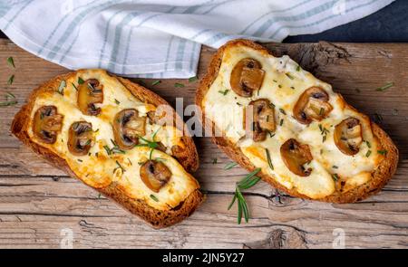 Geröstetes Sandwich aus traditionellem Sauerteig-Brot mit Käse und braunen Pilzen, gewürzt mit Rosmarinkraut auf einem Holzbrett, Draufsicht Stockfoto