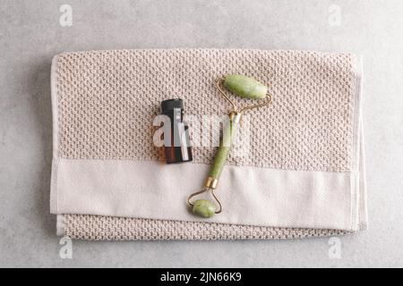 Gesicht Öl und Jade sone gua Sha Massagerolle auf beigefarbenem Handtuch Hintergrund. Beauty- und Hautpflegekonzept, flaches Lay. Stockfoto