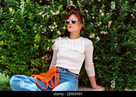 Junge Frau in leichter Sommerkleidung mit wiederverwendbarem Netzbeutel und Sonnenbrille, Sitzbank im Sommerpark vor dem Einkaufen. Nachhaltiger Lebensstil. Stockfoto