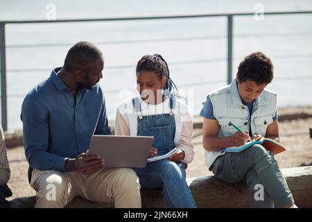 Gruppe lächelnder schwarzer Kinder mit Lehrer, die zusammen einen Laptop während des Unterrichts im Freien in der Sommerschule benutzen Stockfoto