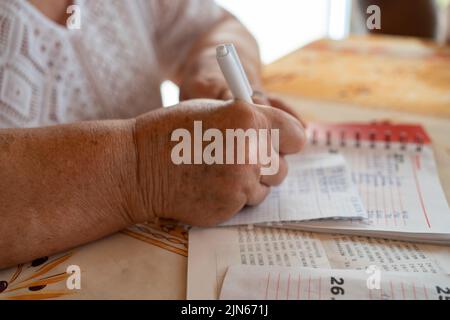 Nahaufnahme der Hände einer älteren kaukasischen Frau, die zu Hause an einem Tisch sitzt und in einem Notizbuch über die Ausgaben Rechenschaft abgibt Stockfoto
