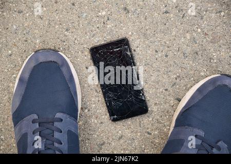 Schwarzes Smartphone mit gebrochenem Display in der Nähe der Füße einer Person auf dem Asphalt. Beschädigung des Telefonbildschirms. Stockfoto