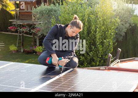 Der junge Mann konzentrierte sich auf die Arbeit, um Solarpanels mit einem elektrischen Schraubendreher auf das Dach zu Schrauben Stockfoto