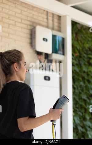Vertikale Seitenansicht einer jungen Frau mit einer Steckdose für Elektroautos auf der Hand und einem Solarsystem im Hintergrund Stockfoto
