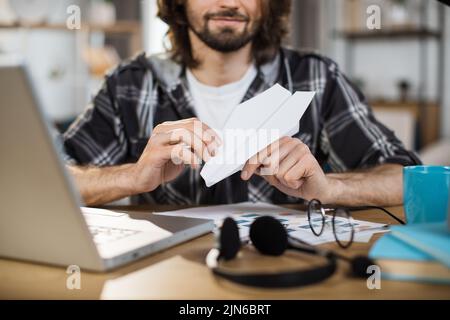 Nahaufnahme eines jungen Mannes, der Papierflugzeug macht, während er am Tisch mit Laptop und Dokumenten sitzt. Gelangweilter Büroangestellter, der Spaß macht und am Arbeitsplatz spielt. Stockfoto